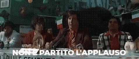 a man in a suit and tie is sitting at a table talking into a microphone while a crowd watches .